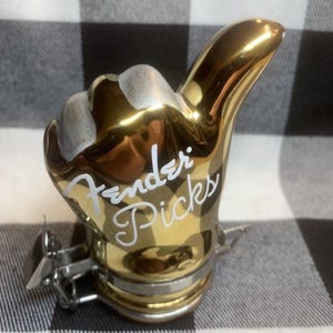 May include: A gold-colored, thumbs-up shaped guitar pick holder with the words "Fender Picks" in white script. The holder has a silver-colored metal base and a clip. The background is a black and white checkered pattern.