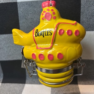 May include: A yellow ceramic ornament shaped like a submarine with the word BEATLES printed in black on the side. The submarine features pink circular accents and a silver metal latch mechanism at the base, suspended by a thin silver string.