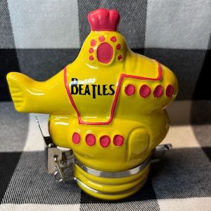 May include: A yellow ceramic jar shaped like a submarine with the word BEATLES printed in black on the side. The submarine features pink accents around the windows and top hatch, and it includes a silver metal wire bail closure mechanism at the base.