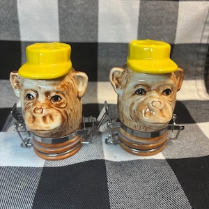May include: A pair of ceramic monkey-shaped salt and pepper shakers. Each shaker features a monkey head with a yellow cap and a brown and tan face. The shakers have a metal clasp and a brown and orange band around the base.