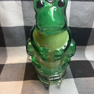 Op de afbeelding: Een groene glazen pot in de vorm van een kikker met een metalen scharniersluiting. De pot heeft grote zwarte ogen en een lichtgroene buik. De achtergrond is een zwart-wit geblokt patroon.