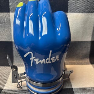 May include: A blue ceramic jar shaped like a fist with yellow fingernails. The word "Fender" is written in white script on the jar. The jar has a metal clasp and a silver band around the base.