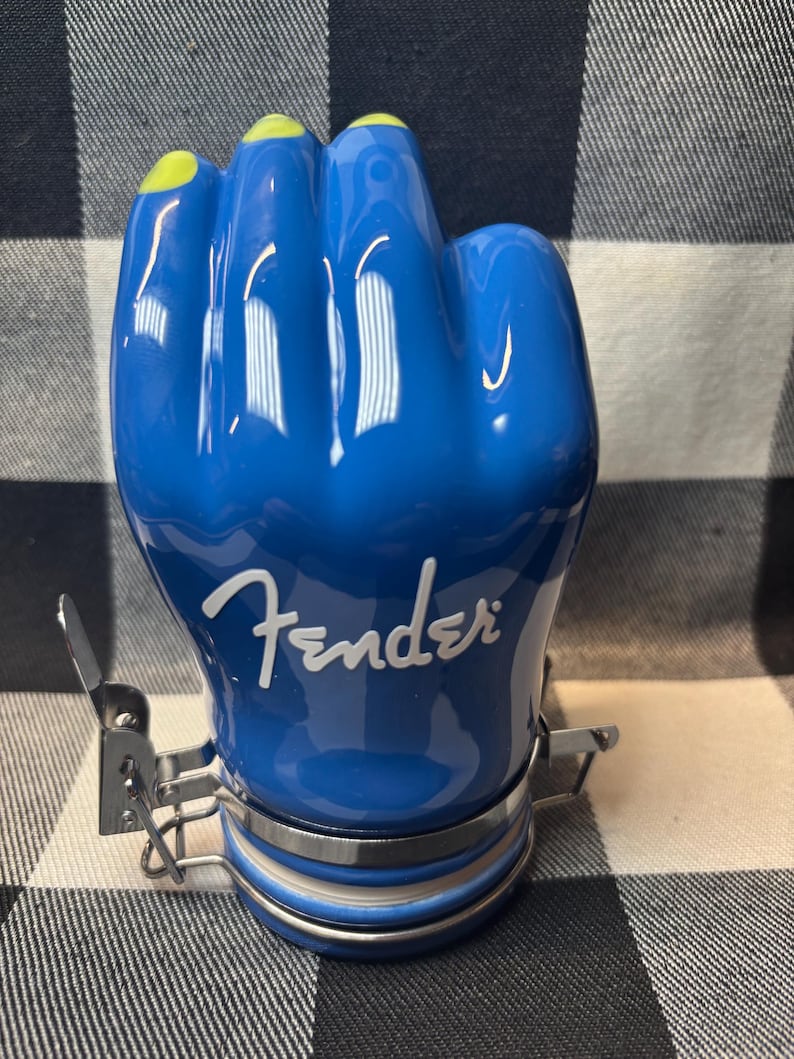 May include: A blue ceramic jar shaped like a fist with yellow fingernails. The word "Fender" is written in white script on the jar. The jar has a metal clasp and a silver band around the base.
