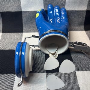 May include: A blue ceramic hand-shaped container with a silver rim and a white interior. The container is open, revealing a coin and several white guitar picks. The lid is blue and white.