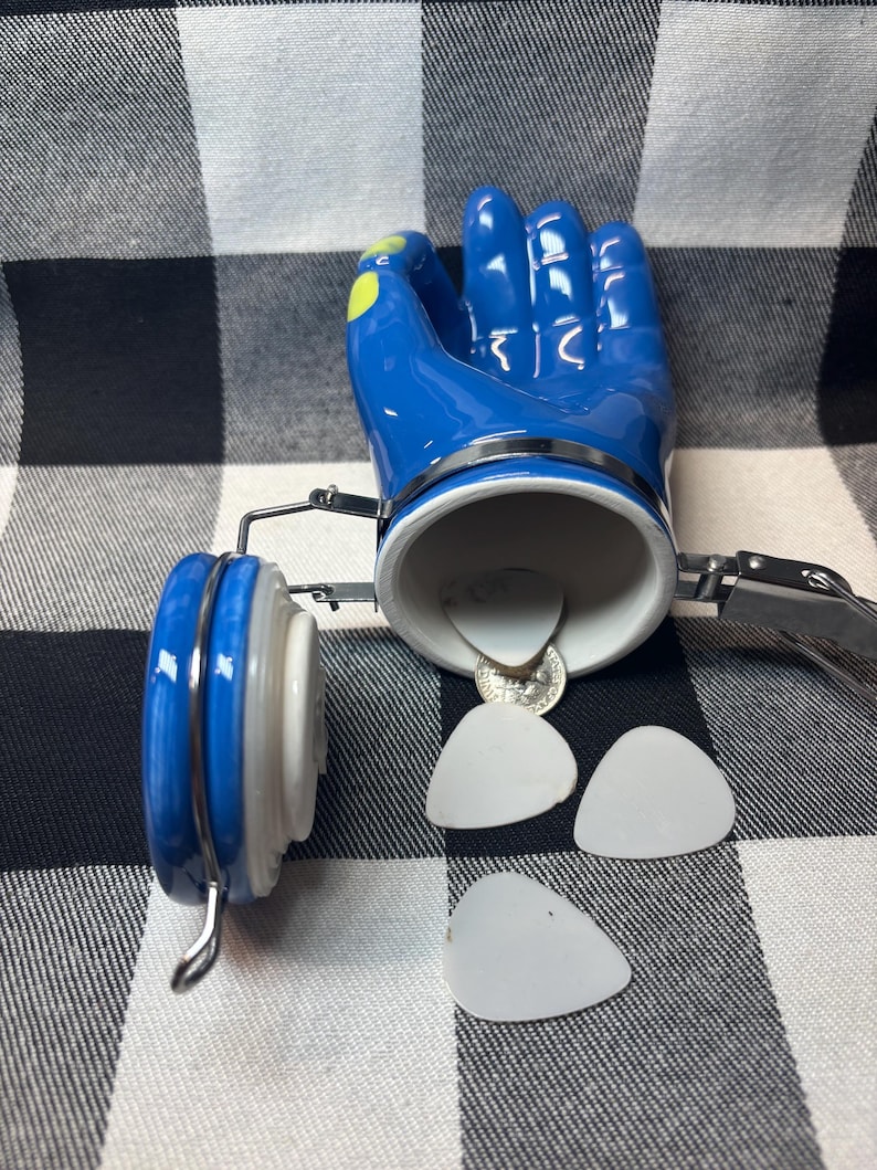 May include: A blue ceramic hand-shaped container with a silver rim and a white interior. The container is open, revealing a coin and several white guitar picks. The lid is blue and white.