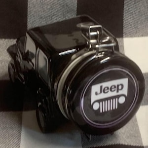 Könnte beinhalten: Ein schwarzes Jeep-Glas mit silbernem Metallverschluss. Das Glas hat die Form eines Oldtimer-Jeeps, mit dem Jeep-Logo prominent auf dem Reserverad. Das Glas steht auf einer karierten Oberfläche.
