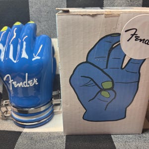 May include: A blue ceramic Fender guitar pick holder shaped like a hand with yellow fingernails. The Fender logo is visible on the hand and on the box. The box features a blue hand graphic with green fingernails.