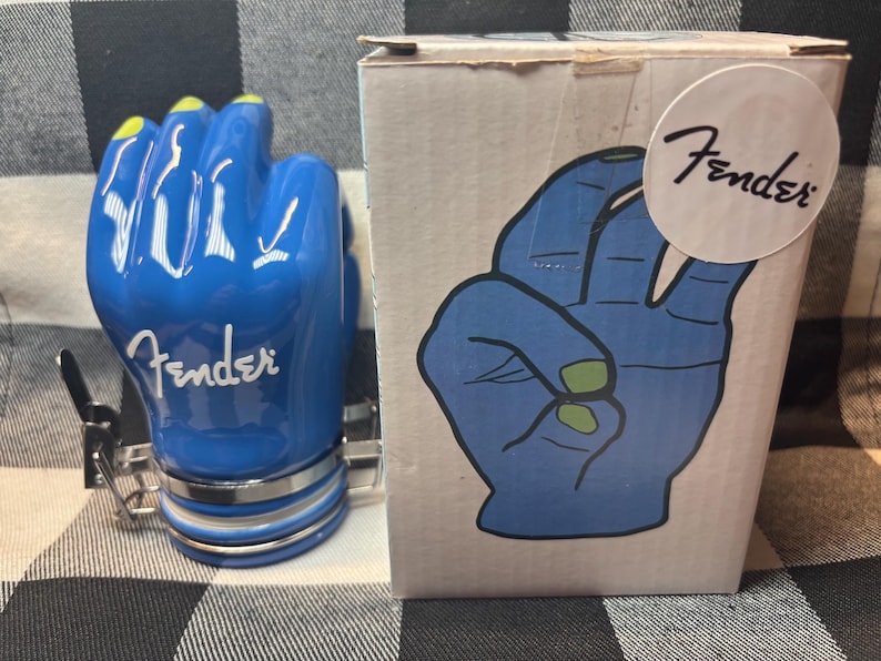 May include: A blue ceramic Fender guitar pick holder shaped like a hand with yellow fingernails. The Fender logo is visible on the hand and on the box. The box features a blue hand graphic with green fingernails.