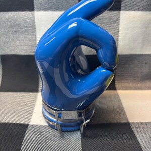 May include: A blue hand sculpture in the 'OK' gesture, with a glossy finish. The fingertips are accented with yellow paint. A silver metal band secures the base of the hand. The sculpture is set against a black and white checkered background.