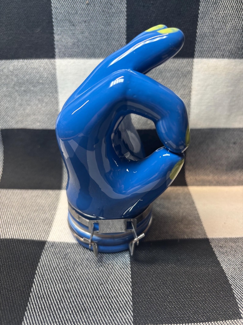 May include: A blue hand sculpture in the 'OK' gesture, with a glossy finish. The fingertips are accented with yellow paint. A silver metal band secures the base of the hand. The sculpture is set against a black and white checkered background.