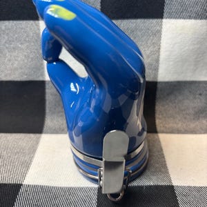 May include: A blue ceramic bottle stopper in the shape of a hand, with yellow accents on the fingertips. The stopper has a silver metal clasp and a silver band around the base. The background is a black and white checkered pattern.