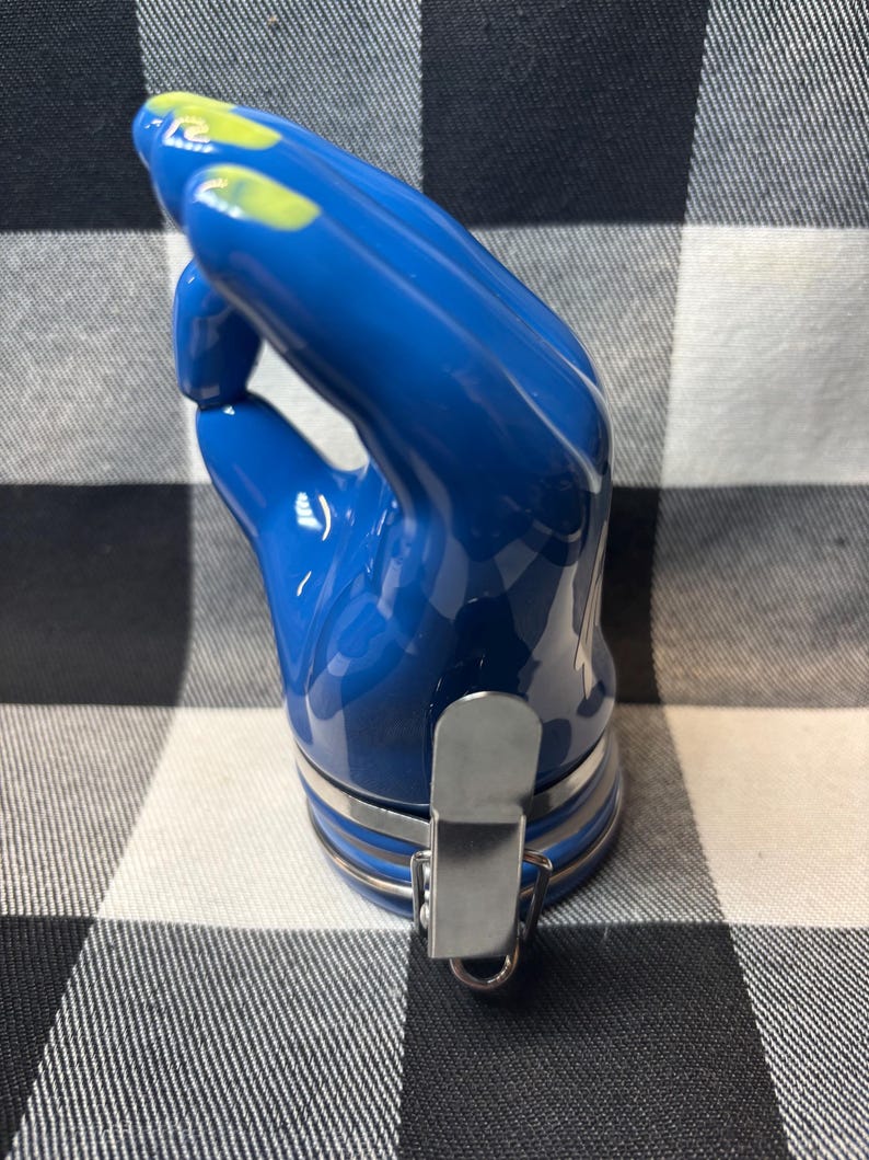 May include: A blue ceramic bottle stopper in the shape of a hand, with yellow accents on the fingertips. The stopper has a silver metal clasp and a silver band around the base. The background is a black and white checkered pattern.
