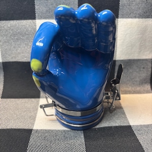 May include: A blue ceramic hand-shaped jar with a silver metal clasp and trim. The jar has a glossy finish and yellow accents on the fingertips. The jar is sitting on a black and white checkered surface.