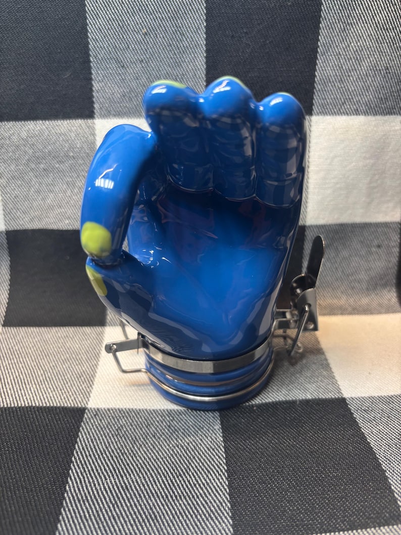 May include: A blue ceramic hand-shaped jar with a silver metal clasp and trim. The jar has a glossy finish and yellow accents on the fingertips. The jar is sitting on a black and white checkered surface.