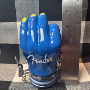 May include: A blue ceramic jar shaped like a hand with yellow fingernails. The word "Fender" is written in white script on the jar. A metal ruler shows the jar is approximately 4 inches tall.