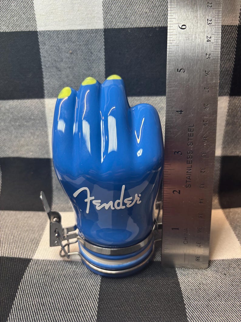 May include: A blue ceramic jar shaped like a hand with yellow fingernails. The word "Fender" is written in white script on the jar. A metal ruler shows the jar is approximately 4 inches tall.