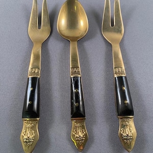 Puede incluir: Un juego de tres utensilios de servir antiguos: dos tenedores y una cuchara. Los utensilios tienen cabezas de metal color oro y mangos negros decorativos con detalles dorados. Los mangos tienen una serie de pequeños puntos dorados.
