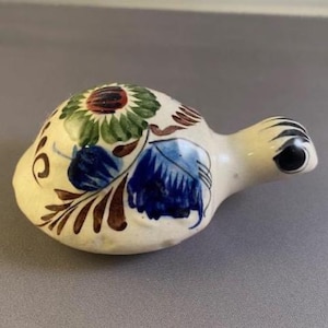 May include: A ceramic bird-shaped ornament with a white base, decorated with floral patterns in blue, green, brown, and red. The bird has a black eye and a curved beak. It is likely a decorative item.