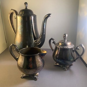 Puede incluir: Un juego de té de tres piezas chapado en plata que incluye una cafetera alta con tapa, una lechera y un azucarero con tapa. Cada pieza presenta una pátina oscura y desgastada con detalles florales ornamentados en las asas y las bases.