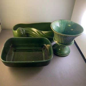 May include: Three green ceramic pieces: a rectangular dish, a long wavy planter, and a pedestal vase. The items are a deep, glossy green, and appear to be vintage or antique. The vase has vertical ribbed detailing.