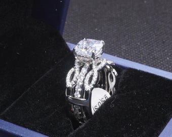 May include: A silver engagement ring set with a large, square-cut clear gemstone and smaller accent stones. The ring is in a blue jewelry box with a black velvet interior. The ring has a unique, intertwined design.