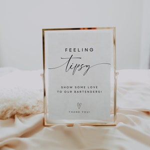 May include: A gold-framed sign with the text "Feeling Tipsy" in cursive script. The sign also includes the text "Show some love to our bartenders!" and a heart symbol. The sign is sitting on a white fabric surface.