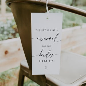 Op de afbeelding: Een wit bord met zwarte tekst die "This row is kindly reserved for the brides family" luidt, met een klein hartje onderaan. Het bord hangt aan een stoel met een wit touwtje.