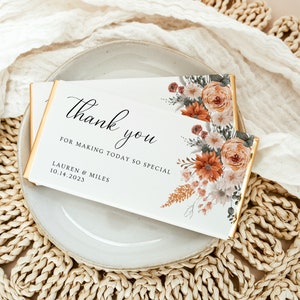 May include: A white chocolate bar with gold foil edges, featuring floral artwork in shades of orange, peach, and green. The text reads "Thank you for making today so special" with a date and names.