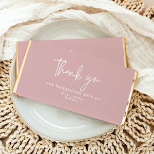 Könnte beinhalten: Zwei rosa Schokoriegel mit goldenen Rändern liegen auf einem grauen Teller, vor einem weißen Tuch und einer gewebten Matte. Die Riegel tragen den Schriftzug "Thank you" in Schreibschrift, sowie den Text "FOR CELEBRATING WITH US!" und Namen und ein Datum.