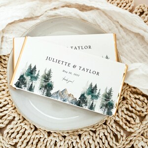 May include: Two rectangular cards with gold edges, featuring a watercolor forest and mountain scene. The cards are inscribed with "JULIETTE & TAYLOR" and "May 20, 2023". The words "Thank you!" are also present.