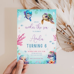 Puede incluir: Una invitación a una fiesta de cumpleaños número 6 con un tema de bajo el mar. La invitación presenta un delfín azul y verde, una tortuga marina verde y un arrecife de coral rosa. El texto de la invitación dice "Únete a nosotros bajo el mar para celebrar que Amelia cumple 6 años".