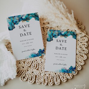 May include: Two save the date cards with blue and gold watercolor designs. The cards feature the text "Save the Date" with the names Harlow & Oliver and the date July 15, 2023. The cards are on a woven placemat.