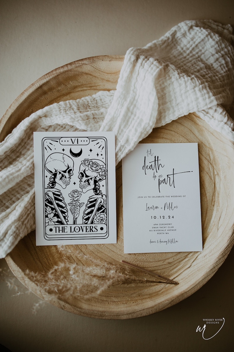 May include: A wedding invitation and a tarot card are displayed on a wooden tray with a white cloth. The tarot card features a black and white illustration of skeletons. The invitation reads "Til death do us part."
