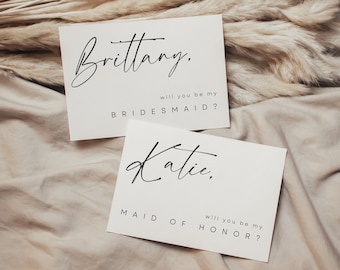 以下が含まれることがあります： 黒い文字が書かれた白いカードが2枚。上のカードには「Brittany. ブライズメイドになってくれますか？」と書いてあります。下のカードには「Katie. ブライズメイドになってくれますか？」と書いてあります。
