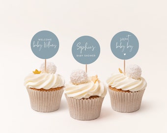 May include: Three cupcakes decorated with white frosting and topped with white sprinkles and a gold star. Each cupcake has a blue circle topper with white text. The toppers read "Welcome Baby Williams", "Sophie's Baby Shower", and "Sweet Baby Boy".