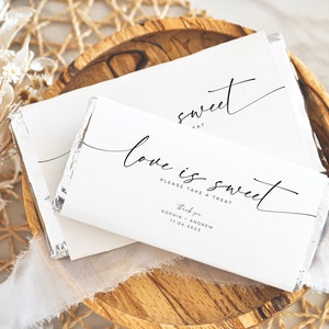May include: White chocolate bar wrapped in silver foil with a white paper label. The label has black calligraphy that reads "Love is sweet" and "Please take a treat". Below the text is "Thank you" and the names "Sophia & Andrew" and the date "17.06.2023".