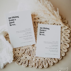 Op de afbeelding: Twee witte huwelijksuitnodigingen met zwarte tekst. De tekst luidt "nothing fancy just love". De uitnodigingen bevatten de datum, tijd en locatie van de bruiloft. De uitnodigingen liggen op een geweven placemat.