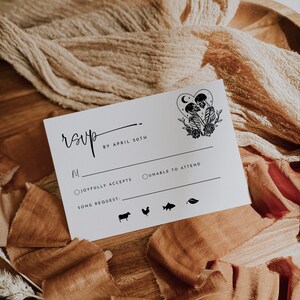 May include: A white RSVP card with black text and a black and white illustration of two skeletons holding hands. The text reads "RSVP by April 20th". There are two radio buttons for guests to select: "Joyfully Accepts" and "Unable to Attend". There is a space for a song request and a row of icons for guests to choose from.