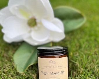 May include: A brown glass jar candle with a black lid, labeled "Sugar Magnolia" with scent notes. A large white magnolia flower and green leaves are in the background. The candle is 7 oz.
