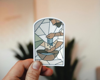 May include: A sticker with a stained-glass style illustration. The design depicts hands pouring water into a bowl. The sticker has a white border and is held by a hand.