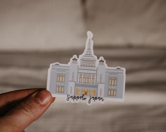 May include: A sticker featuring a gray building with a white roof and windows. The building is labeled "Sapporo Japan".