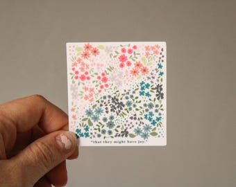 May include: Square sticker with a floral design in pink, peach, gray, blue, and green. The sticker has the text "that they might have joy." The sticker is held by a hand.
