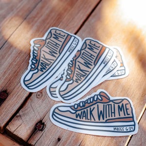 May include: Several stickers featuring a sneaker design with the words "WALK WITH ME" in a stylized font. The stickers are in shades of tan, white, and blue, with the text "MOSES 6:34". The stickers are on a wooden surface.