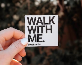 May include: A white square sticker with the words "WALK WITH ME." in bold, black capital letters. Below the text is "MOSES 6:34" in smaller, black text. The sticker is held by a hand with white nail polish.
