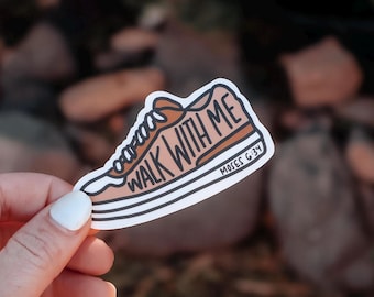 May include: A sticker in the shape of a sneaker with the words "WALK WITH ME" in a brown font. The shoe is tan with white and black stripes on the sole. The text "MOSES G:34" is also on the sticker.