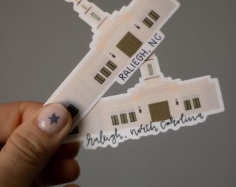 May include: Two stickers featuring a detailed illustration of a building, likely a government building or landmark. The stickers are white with brown and black accents, and the text "RALEIGH, NC" and "raleigh, north carolina" is visible.