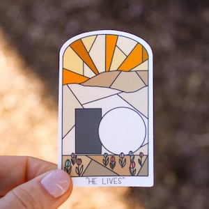 May include: A sticker with a stained glass style illustration of a sunrise over a stone tomb with the text "HE LIVES".