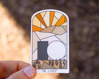 May include: A sticker with a stained glass style illustration of a sunrise over a stone tomb with the text "HE LIVES".
