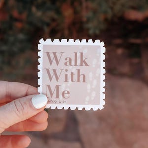 May include: A square sticker with a light pink background and the words "Walk With Me" in a soft brown font. The sticker has a white border with a postage stamp edge. Small footprints are printed on the background.