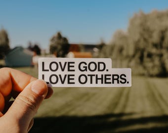 May include: A white sticker with black text that reads "Love God. Love Others." The sticker is being held by a hand against a blurred background of green grass and trees.